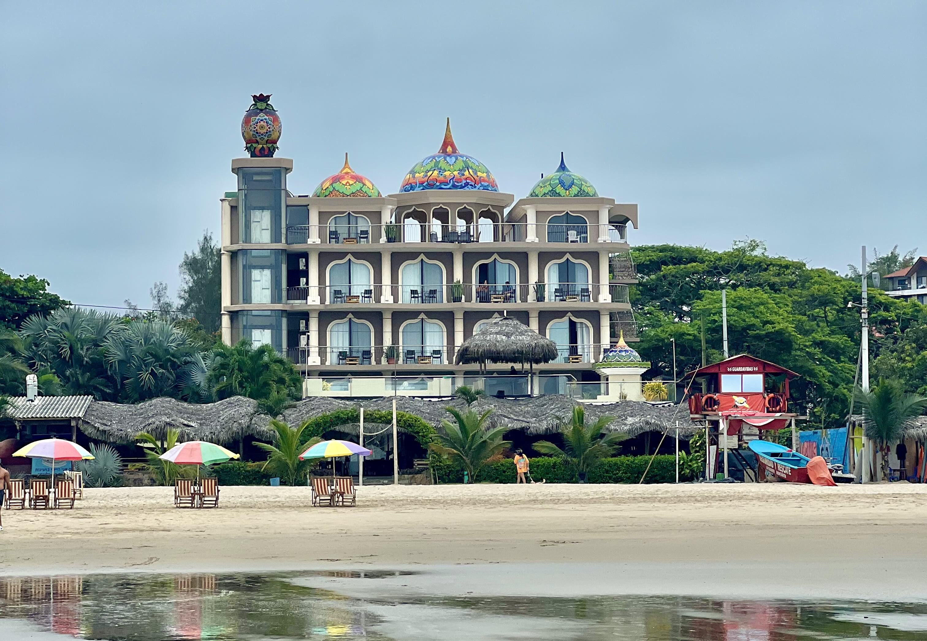 Colorful domes top an eclectic beachfront Dharma Beach building with a large owl statue, flanked by palm-thatched huts and colorful beach umbrellas on the sandy shores of Montañita Beach.