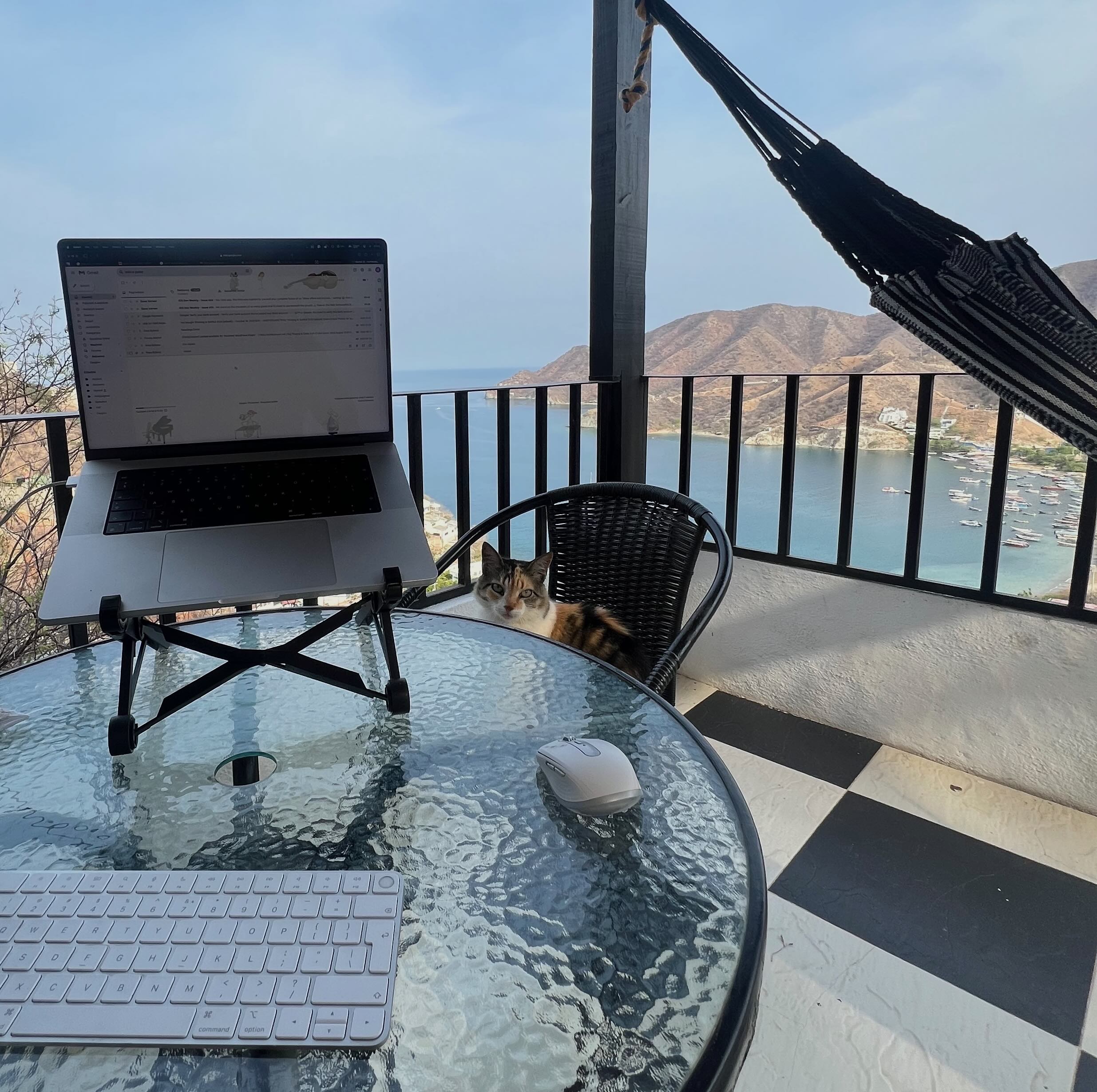 A laptop on a stand sits on a glass table with a keyboard and mouse, overlooking a coastal view of Taganga, Colombia. Raúl is sitting on a chair next to the table, and a hammock is hanging nearby.