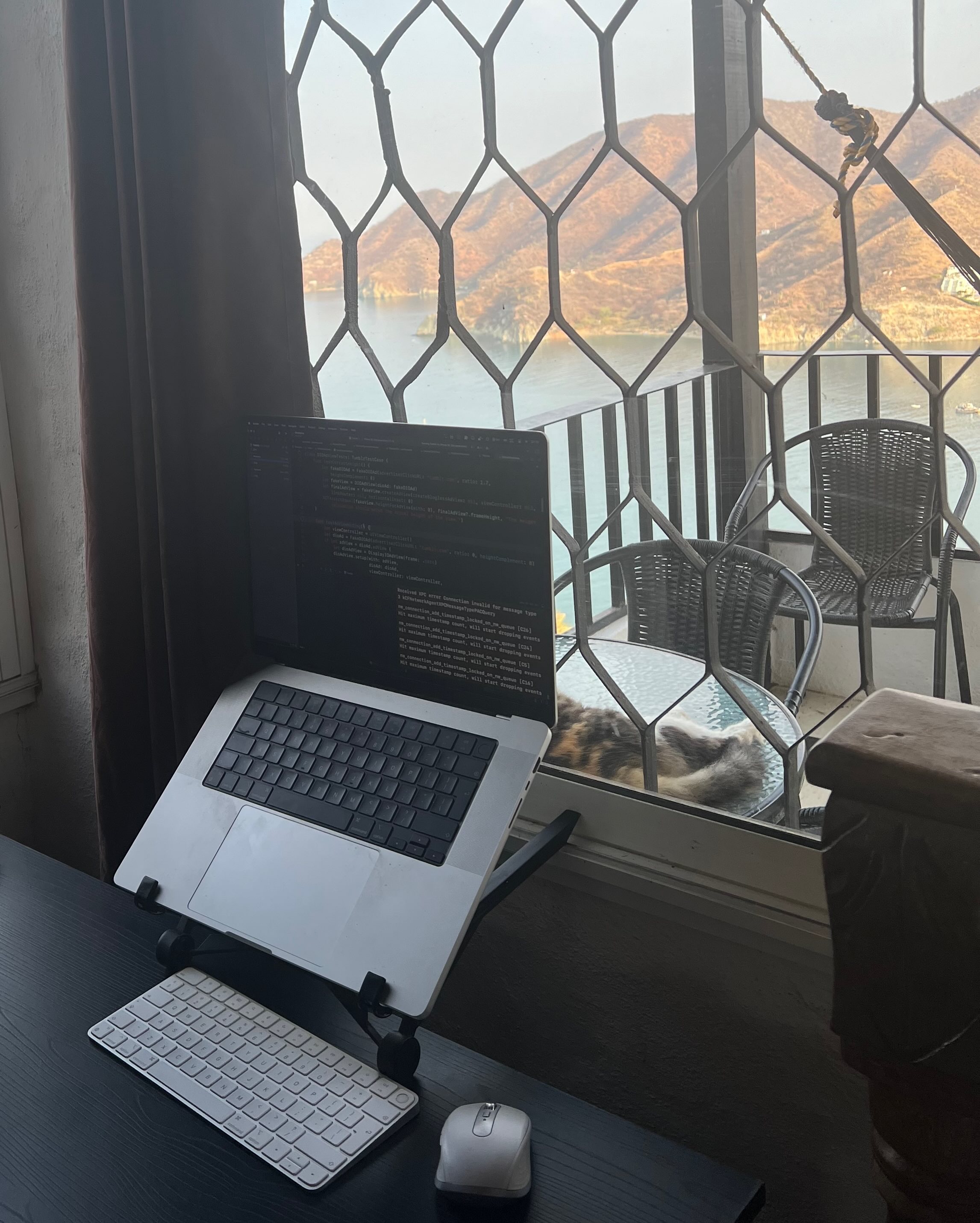 A laptop on a stand sits on a desk with a keyboard and mouse, positioned next to a window with a metal grill. The window overlooks a coastal view of Taganga, Colombia, with mountains in the background and Raúl lounging outside on a chair.