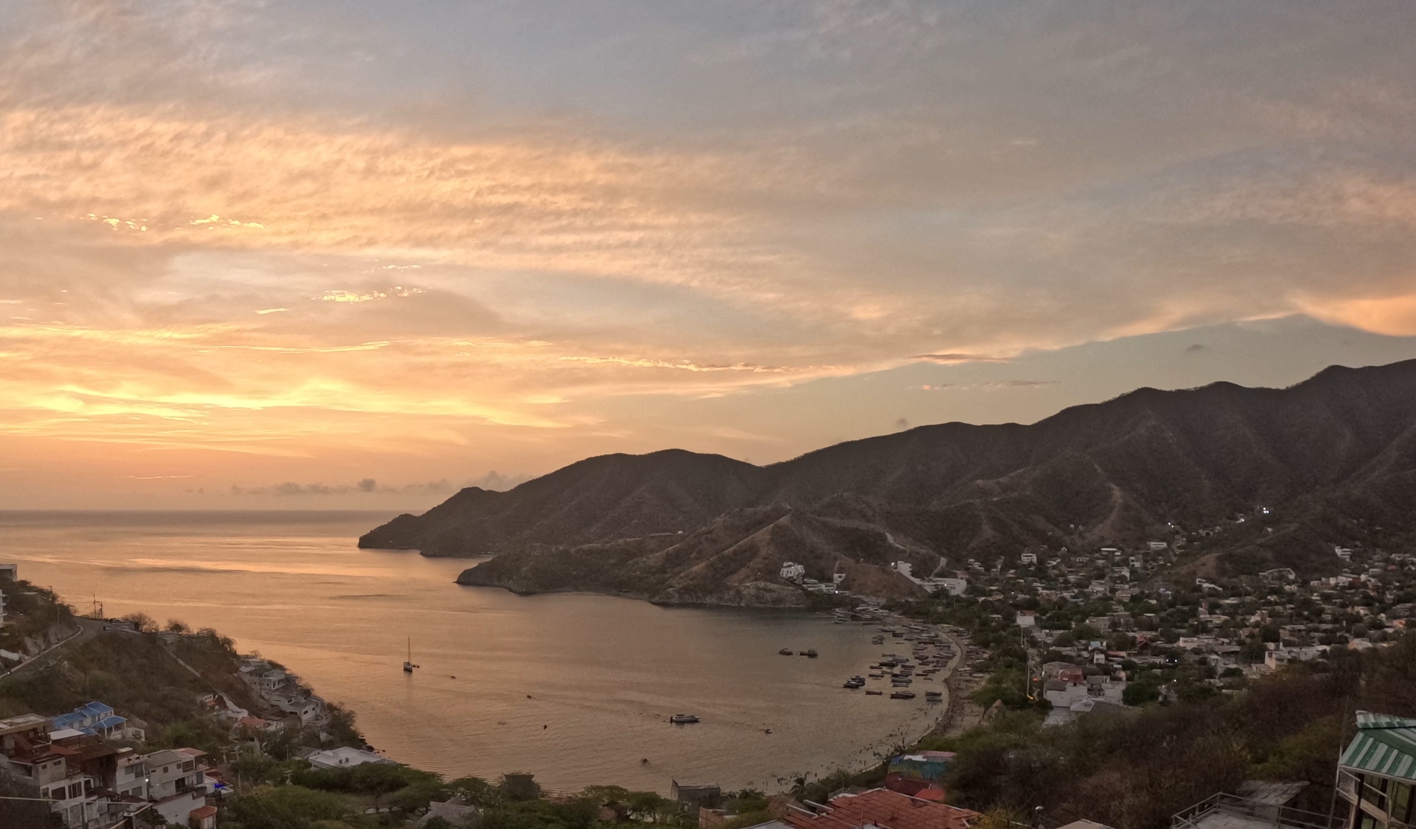 A scenic view of Taganga, Colombia, at sunset, with a golden sky, coastal mountains, and boats dotting the bay, surrounded by hillside houses.