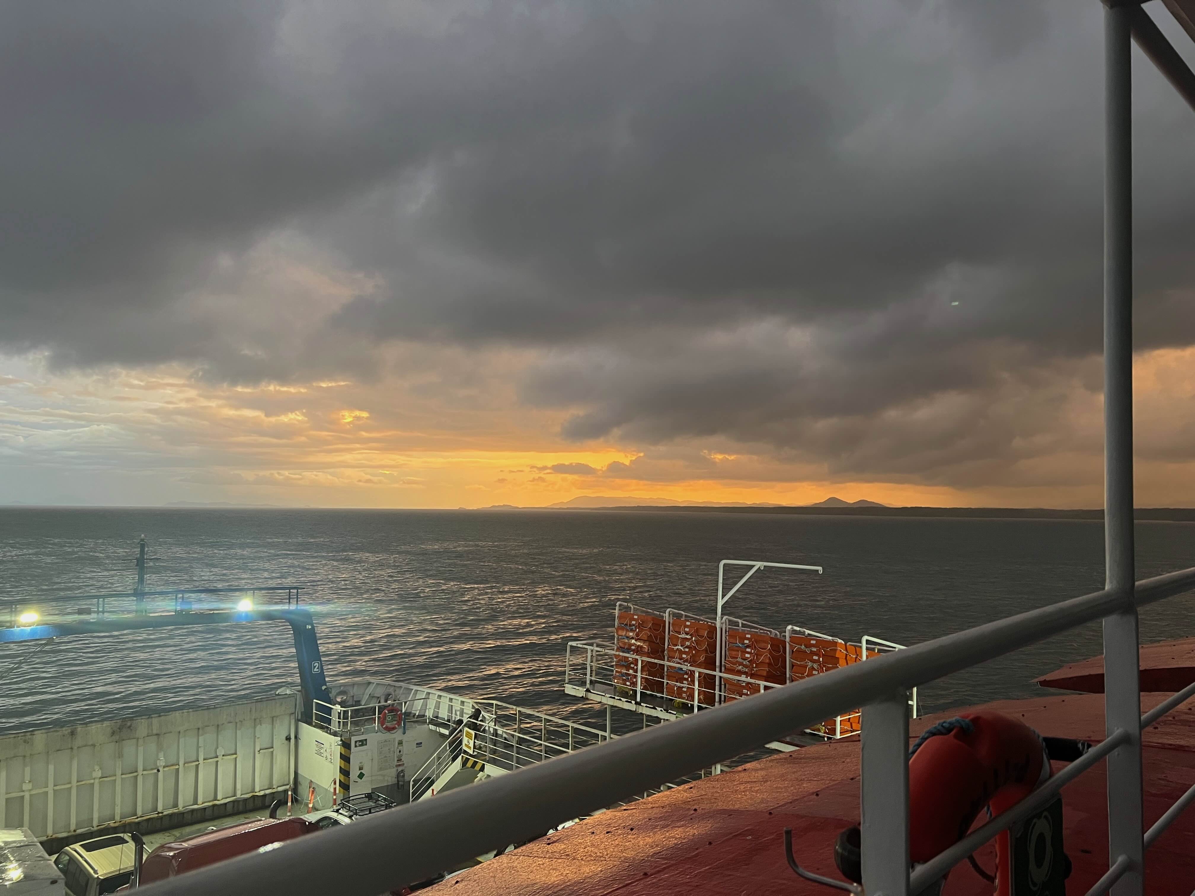 View from a ferry deck showing a cloudy sunset over the ocean, with a silhouette of Santa Teresa far away.