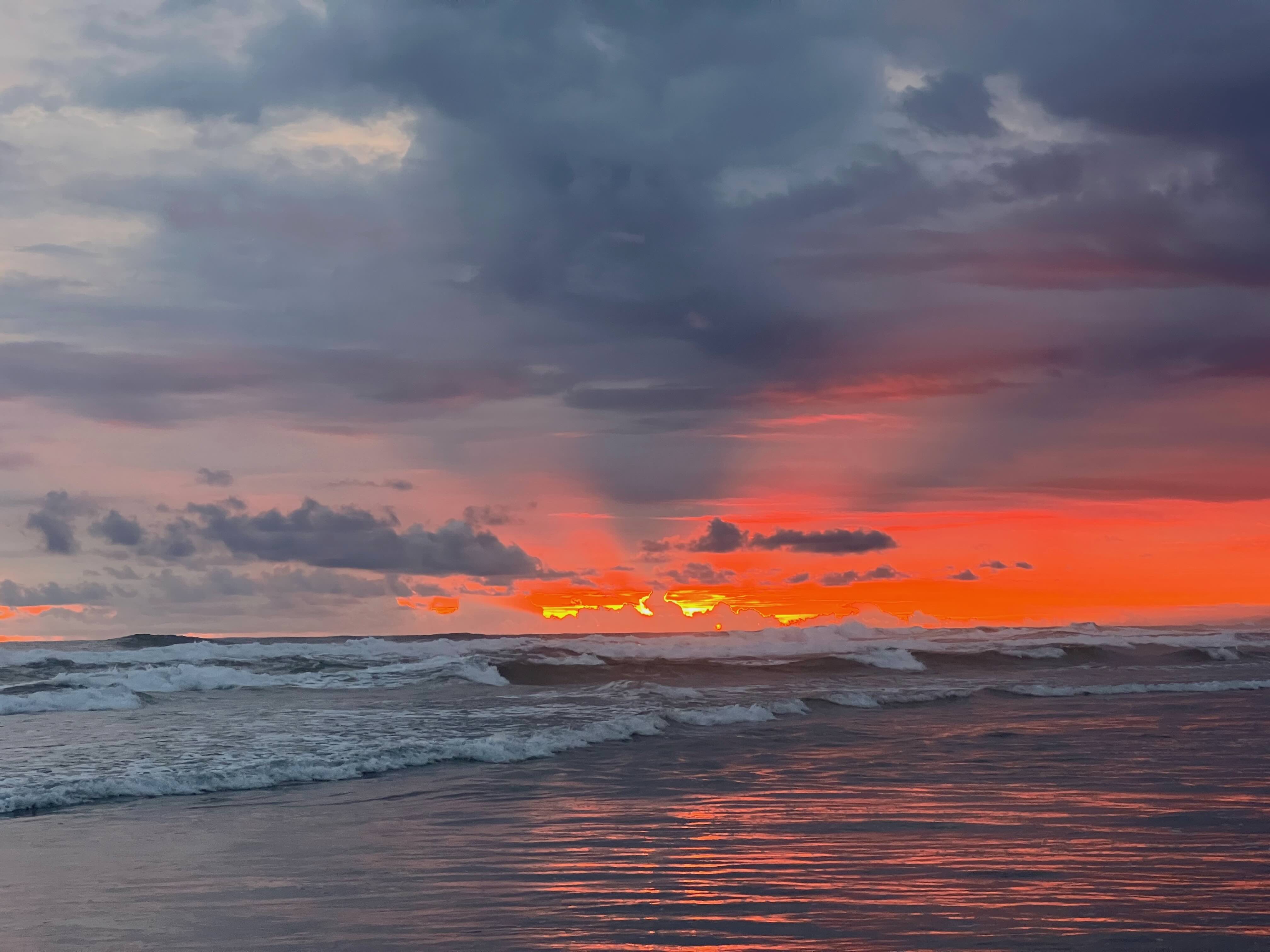 Sunset over the ocean with vibrant orange and red skies reflecting on the water, waves gently crashing on the shore.