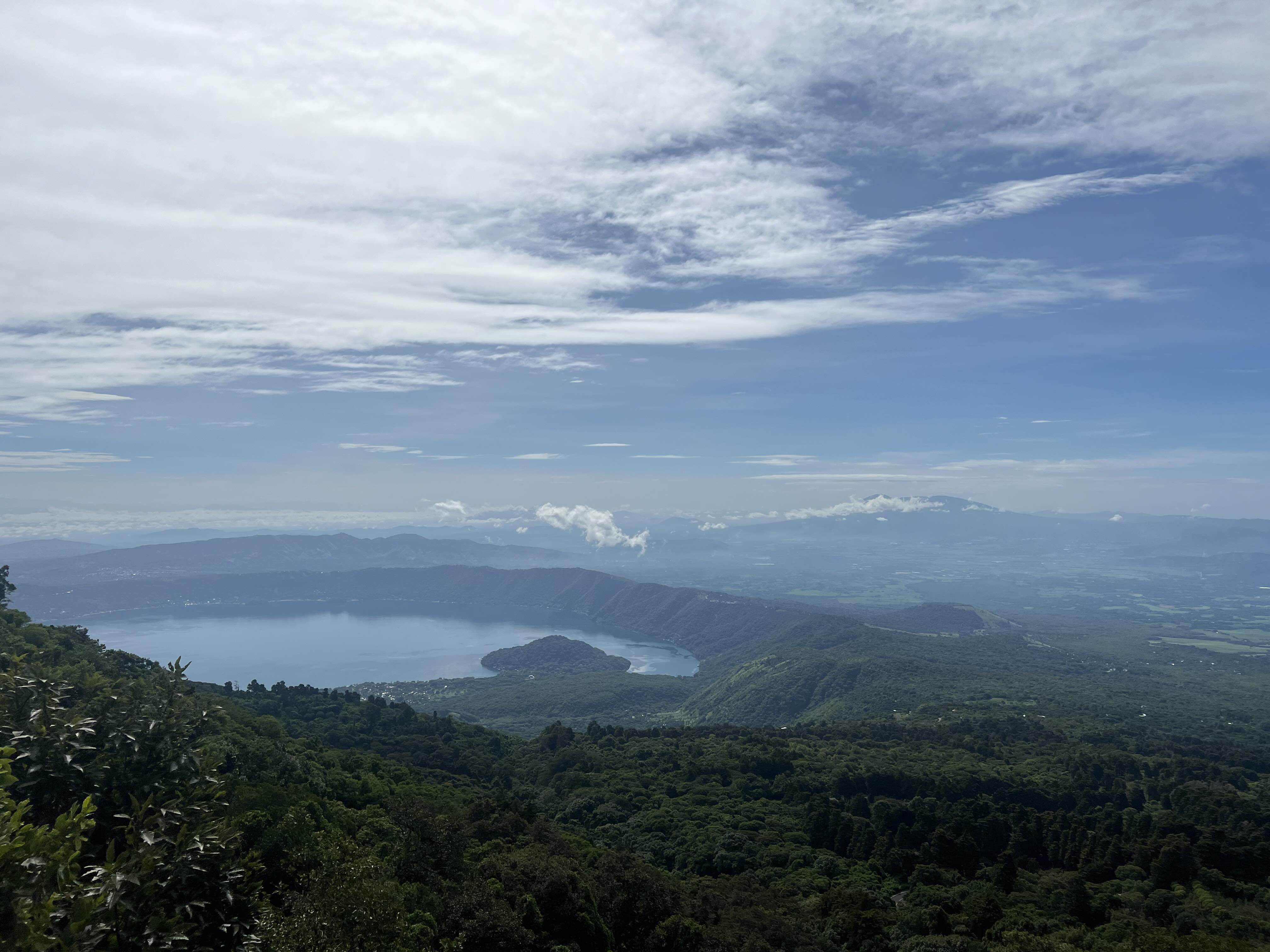 Stunning aerial view of Lake Coatepeque surrounded by lush greenery, seen from the Santa Ana volcano, under a vast, partly cloudy sky.