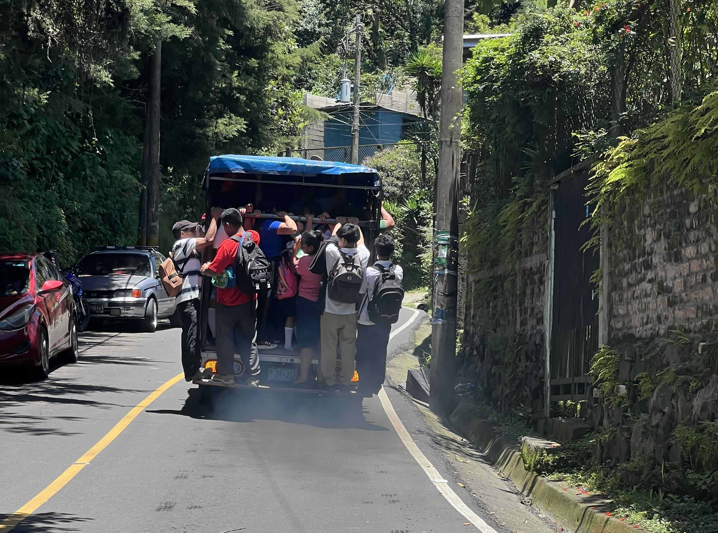 A crowded open-back truck filled with people standing and hanging onto the rear, driving down a narrow road surrounded by greenery