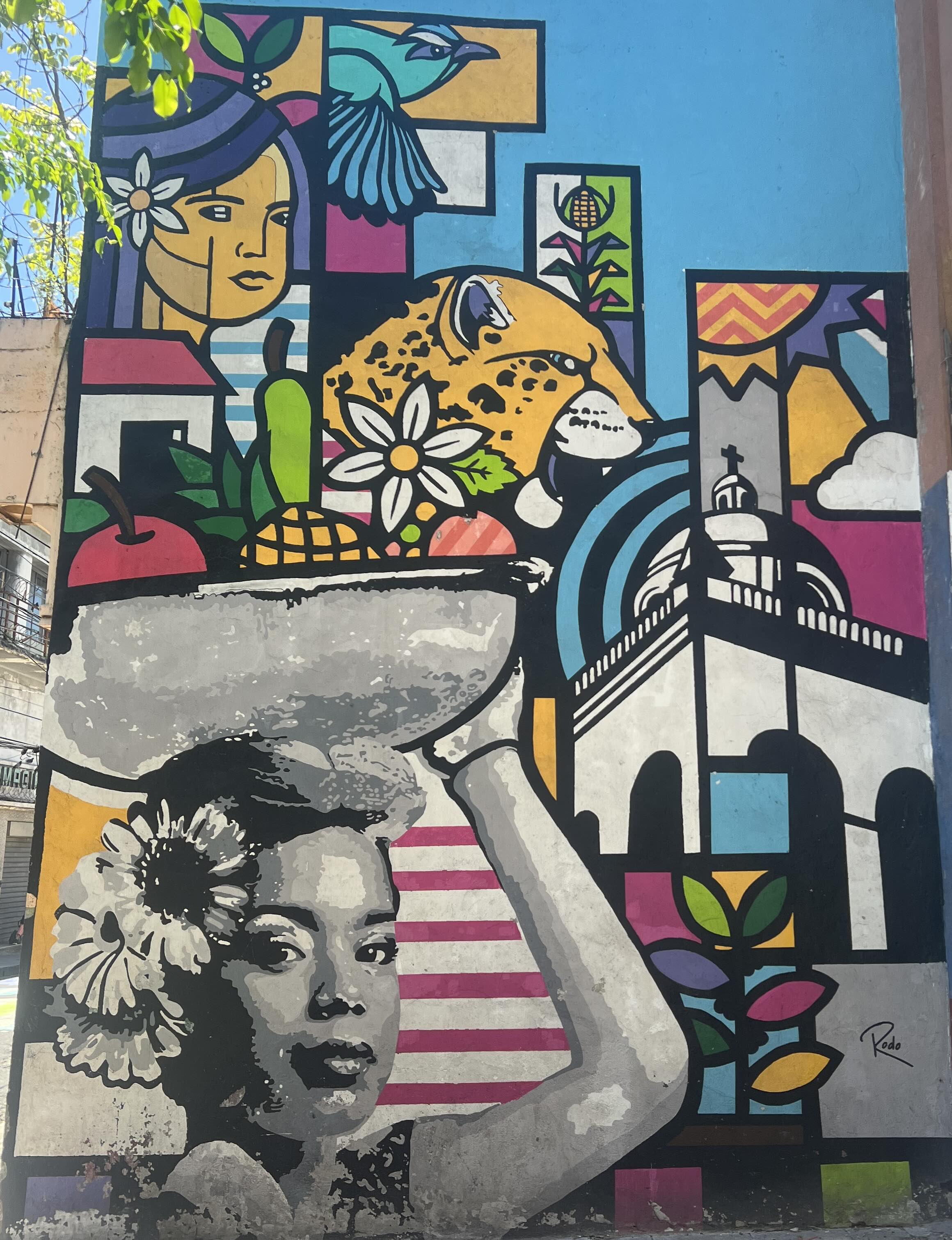 A vibrant mural featuring a woman with a flower in her hair carrying a basket, along with depictions of a jaguar, tropical plants, a bird, and architectural elements, symbolizing the cultural heritage of El Salvador.