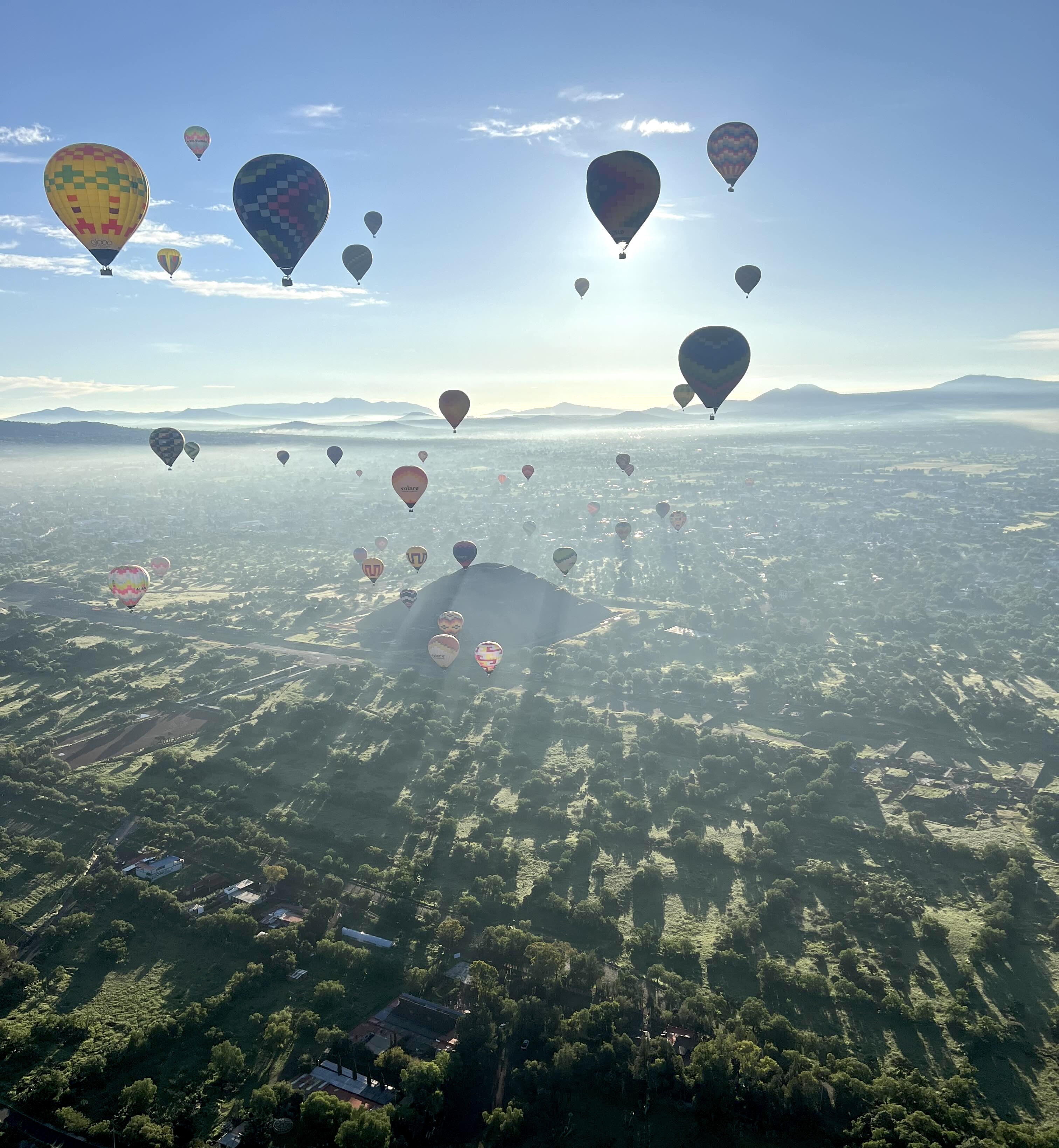 A scenic view of hot air balloons floating over the ancient site of Teotihuacán, with sunlight casting shadows over the lush green landscape and pyramids below.