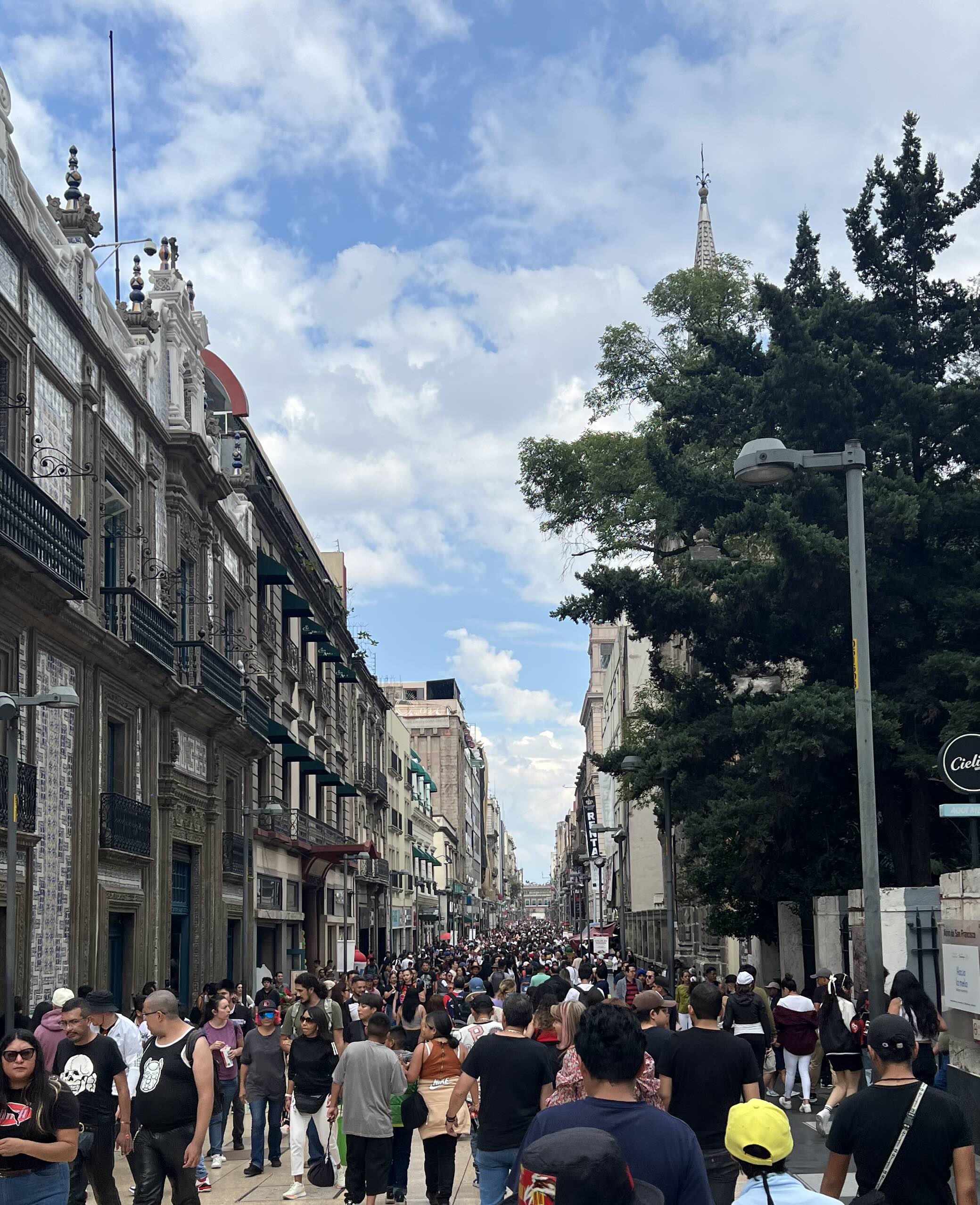 A bustling pedestrian street in Mexico City’s historic center, filled with people walking between historic buildings under a partly cloudy sky.