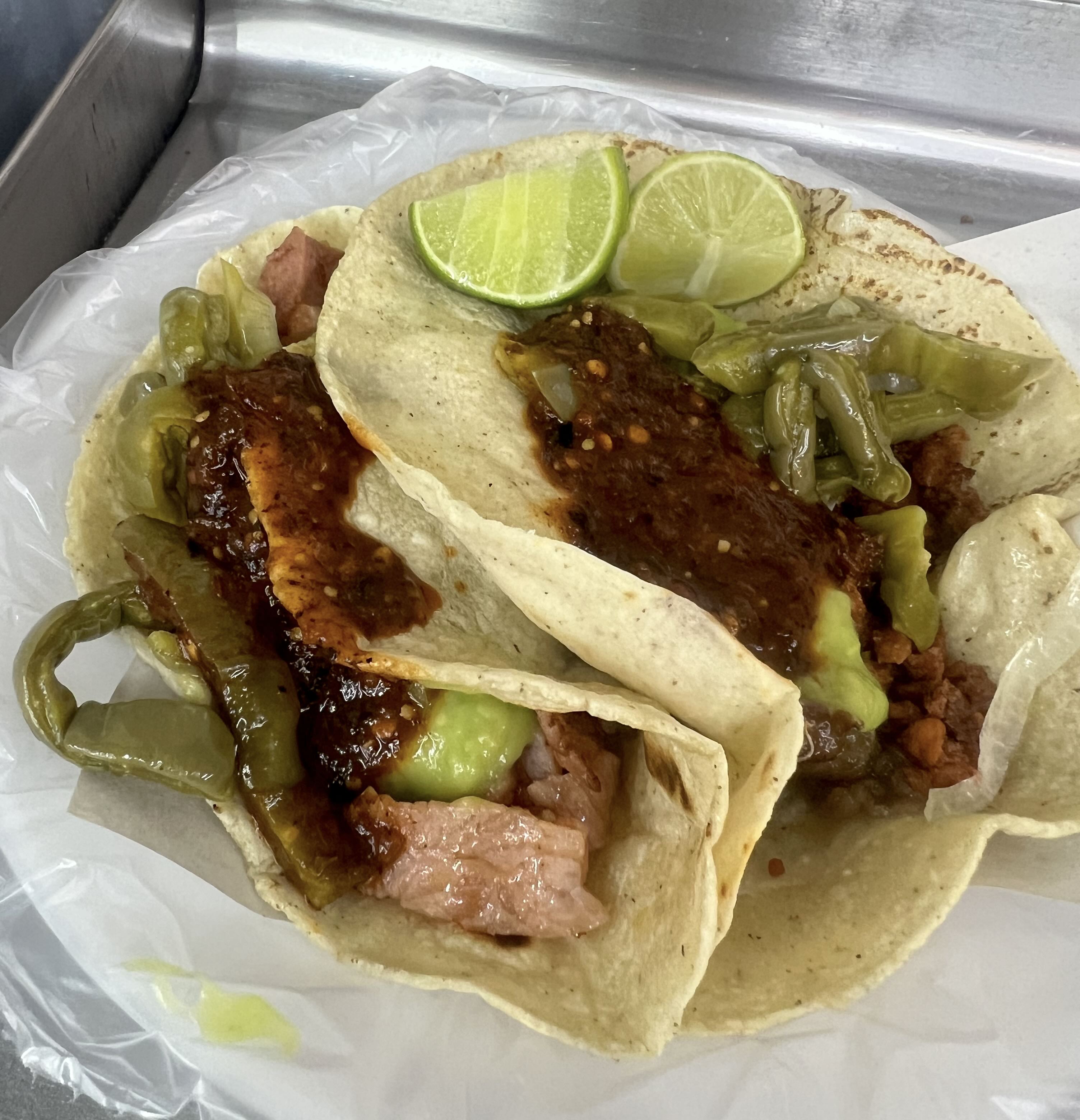 Two tacos topped with salsa and nopales. The taco on the left is taco de chuleta, and the taco on the right is campechano. Lime wedges sit on top for garnish