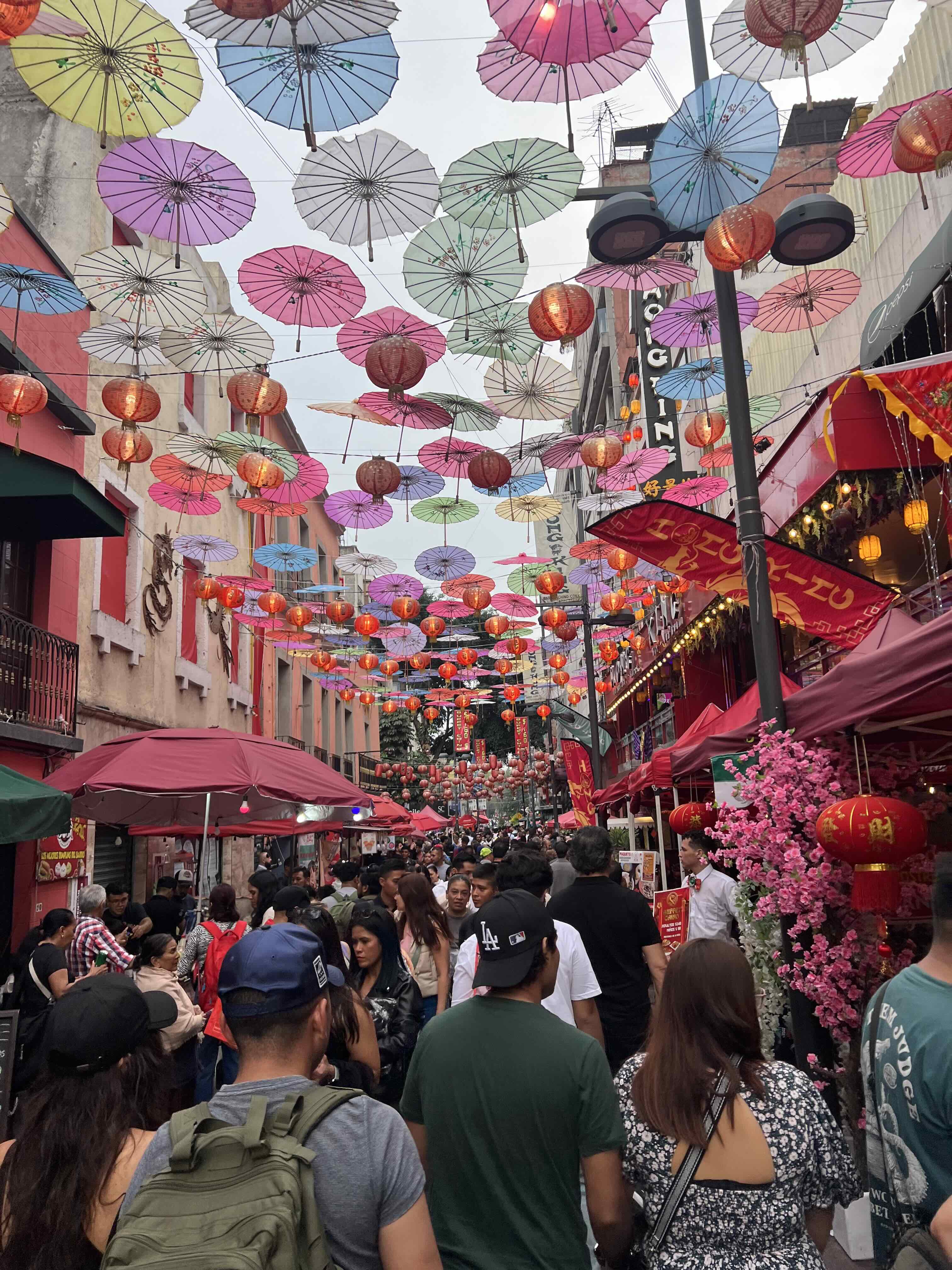 A festive street in Barrio Chino with colorful umbrellas and red lanterns hanging above a crowd.