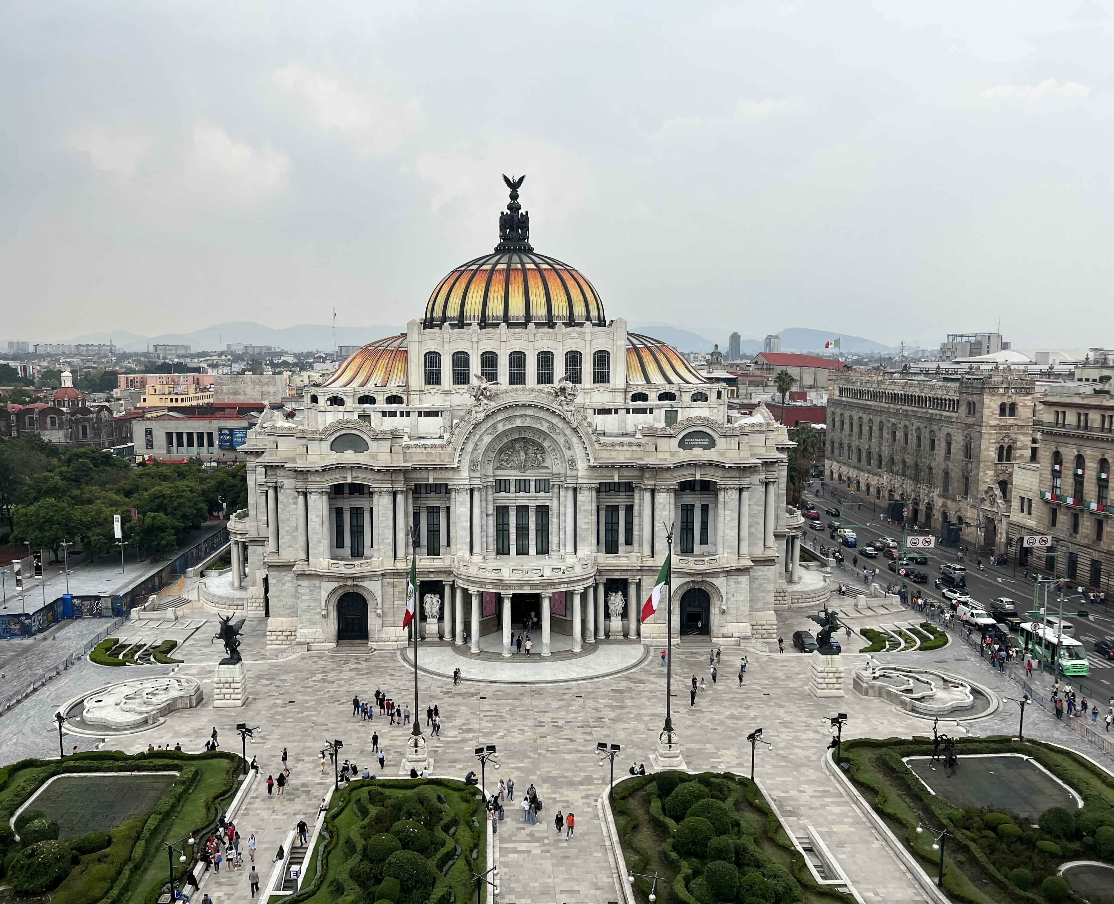 View of Palacio de Bellas Artes, a grand white building with a colorful domed roof, surrounded by a large plaza