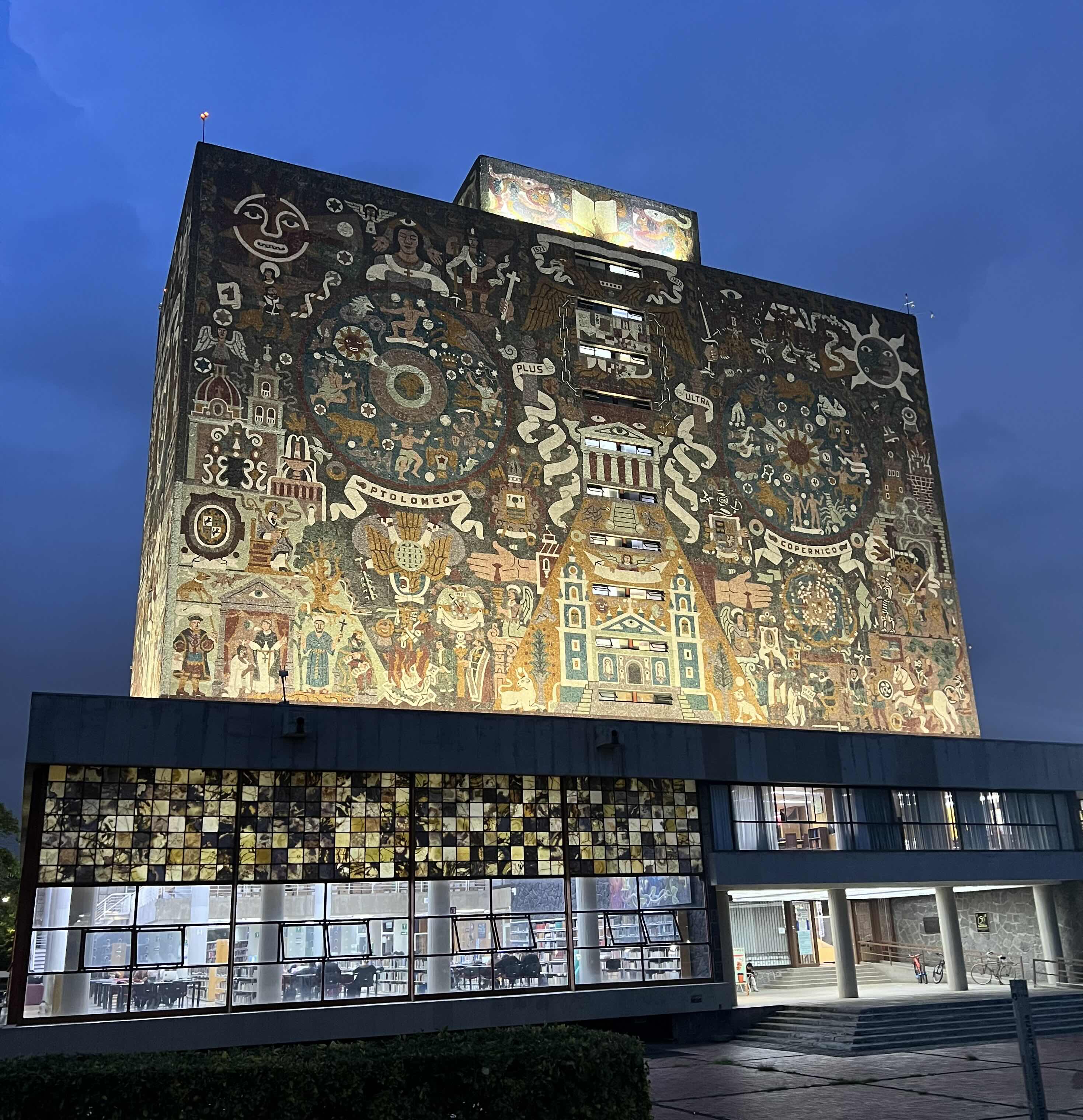 The iconic Central Library at UNAM in Mexico City, featuring its illuminated mural filled with intricate symbols and historic imagery against a twilight sky.